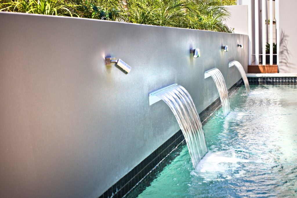 Three water spouts cascade into a pool from a wall. White tiles line the pool's edge, and green plants are visible behind the wall, offering natural privacy solutions.