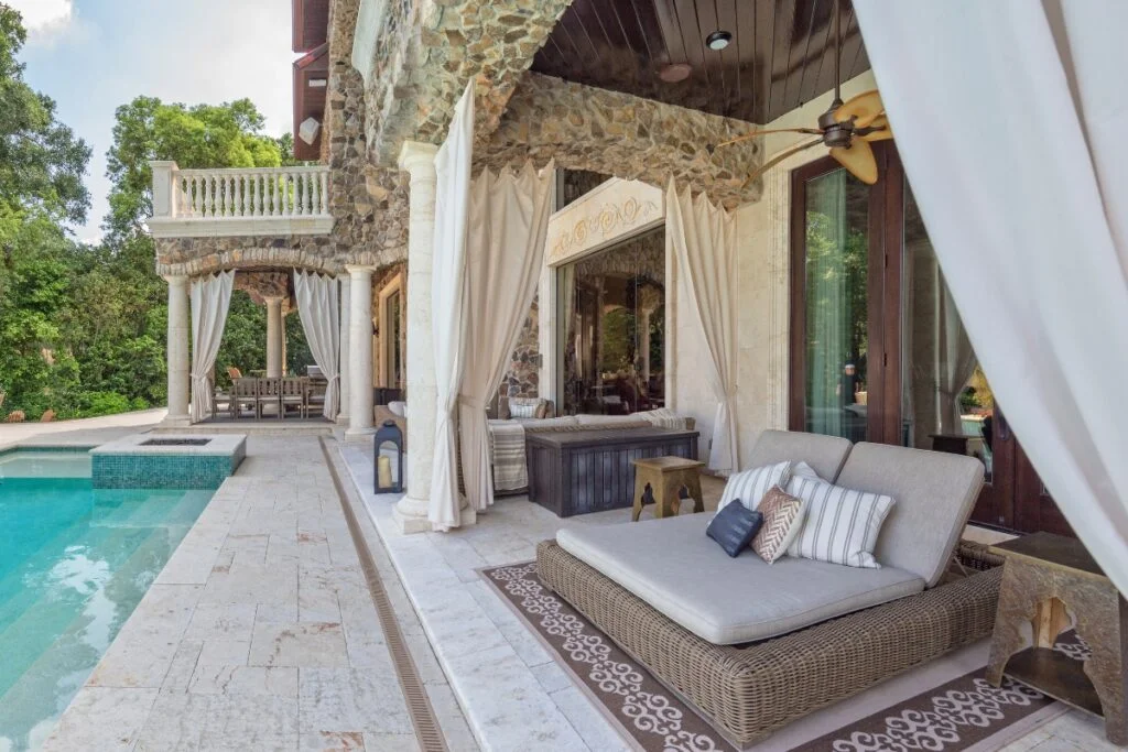 Luxury poolside patio with stone facades, curtains for privacy solutions, lounge chairs, a ceiling fan, and ornamental details. A serene pool is adjacent, partially visible on the left.