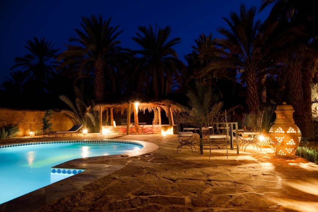 A nighttime view of a poolside area with ambient lighting, surrounded by palm trees. Patio seating, a lit gazebo, and a decorative lantern create a serene atmosphere enhanced by privacy solutions that ensure an intimate getaway.