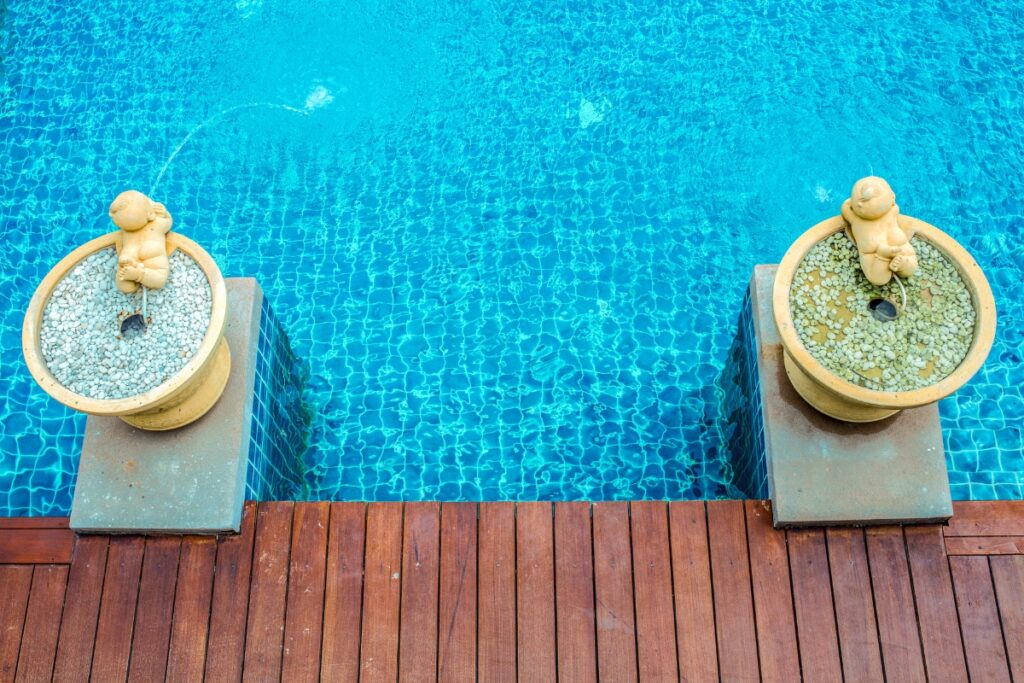 Two child-shaped fountains spout water into a clear blue swimming pool, bordered by a wooden deck with integrated privacy solutions.