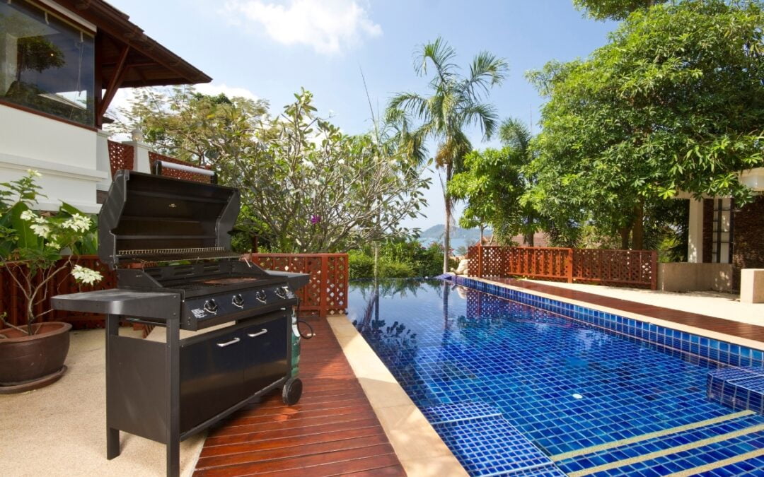 A black barbecue grill is placed on a wooden deck beside a blue-tiled swimming pool in a tropical outdoor setting, showcasing elegant outdoor kitchen designs.