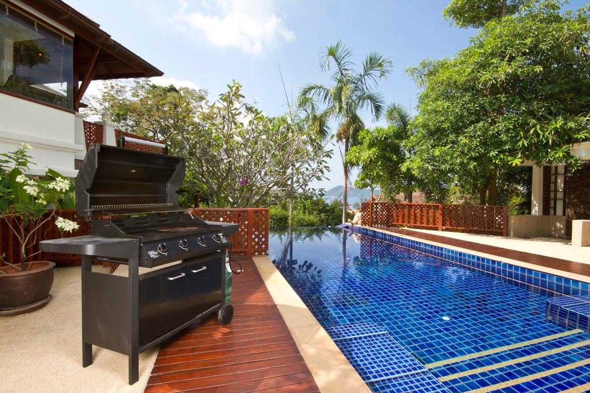 A black barbecue grill with an open lid stands next to a blue-tiled swimming pool in a backyard surrounded by trees and plants, showcasing stylish outdoor kitchen designs.