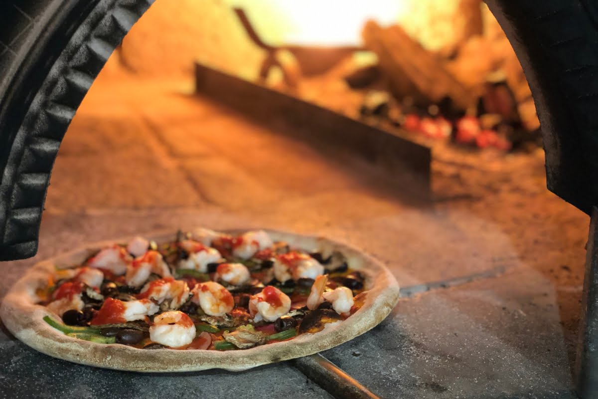 A wood-fired pizza with various toppings is cooking on a stone surface inside a beautifully crafted outdoor kitchen design.