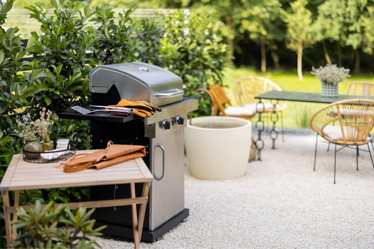 A stainless steel gas grill with kitchen gloves and grilling utensils stands on a patio next to an outdoor dining table and chairs, showcasing one of the finest outdoor kitchen designs surrounded by lush greenery.