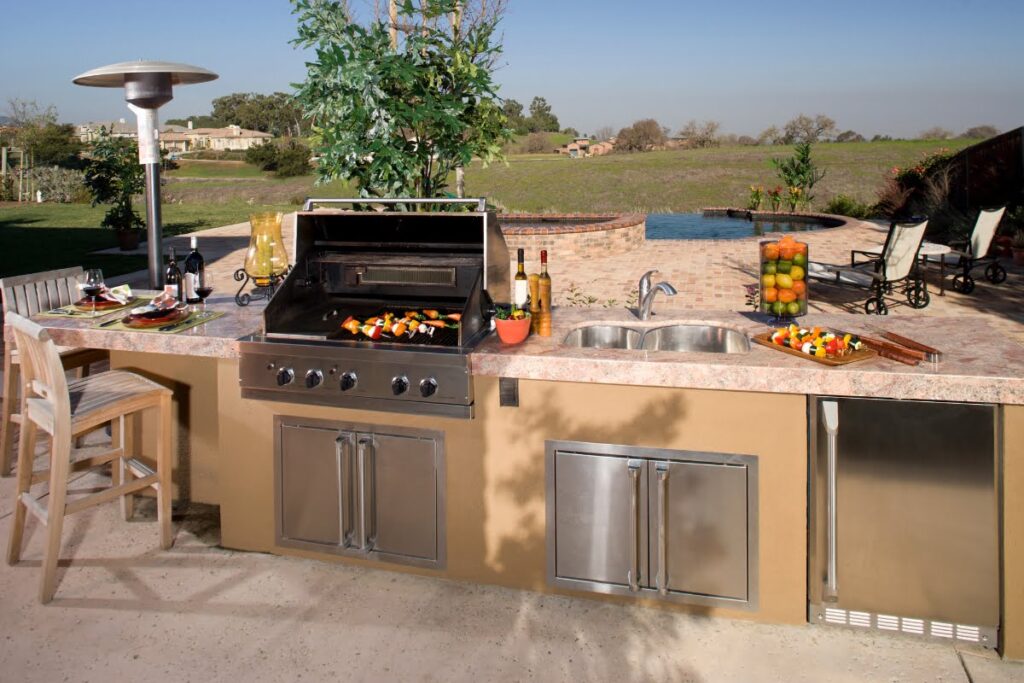 Outdoor kitchens designs feature a built-in grill, sink, refrigerator, and counter seating with food on the countertop, all overlooking a backyard pool and open landscape.