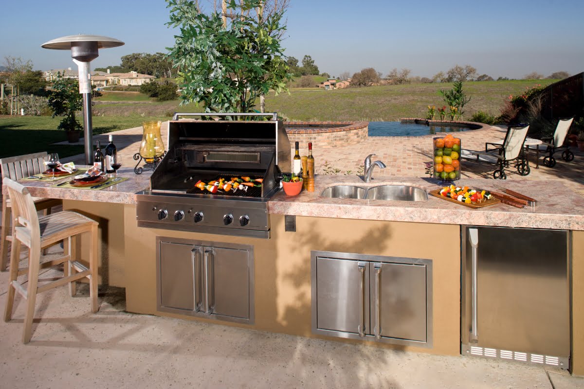 Outdoor kitchen designs featuring a grill, sink, and ample counter space for food and drinks. The area includes a bar stool and is beautifully surrounded by a landscaped yard with a pool in the background.