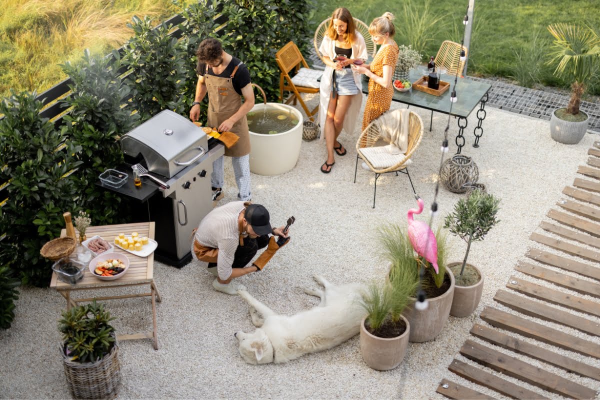 A group of people, two grilling at a barbecue, two seated and using a phone, and a large white dog lying on the ground in an outdoor patio with plants and a pink flamingo ornament—a perfect setting to get inspired by outdoor kitchen designs.