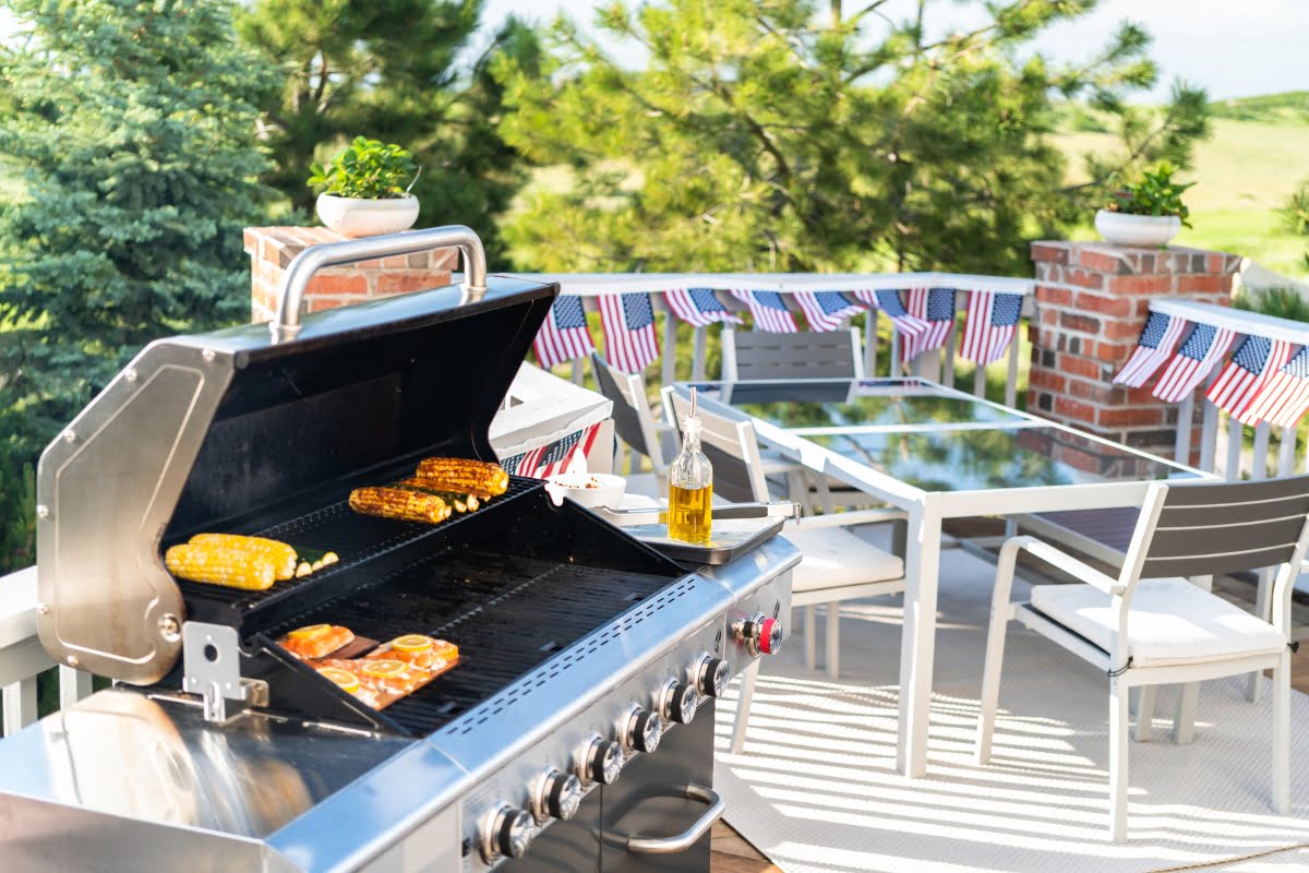 Outdoor deck with innovative outdoor kitchen designs, featuring a grill cooking corn and meat, a glass table with chairs, and American flag decorations.