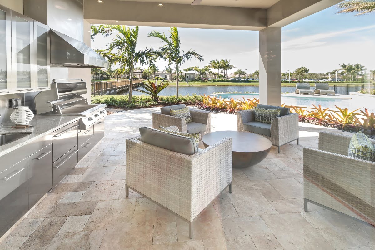 A covered outdoor patio featuring one of the most stylish outdoor kitchen designs, complete with a seating arrangement and a round table, overlooks a pool, palm trees, and a lake under a clear sky.