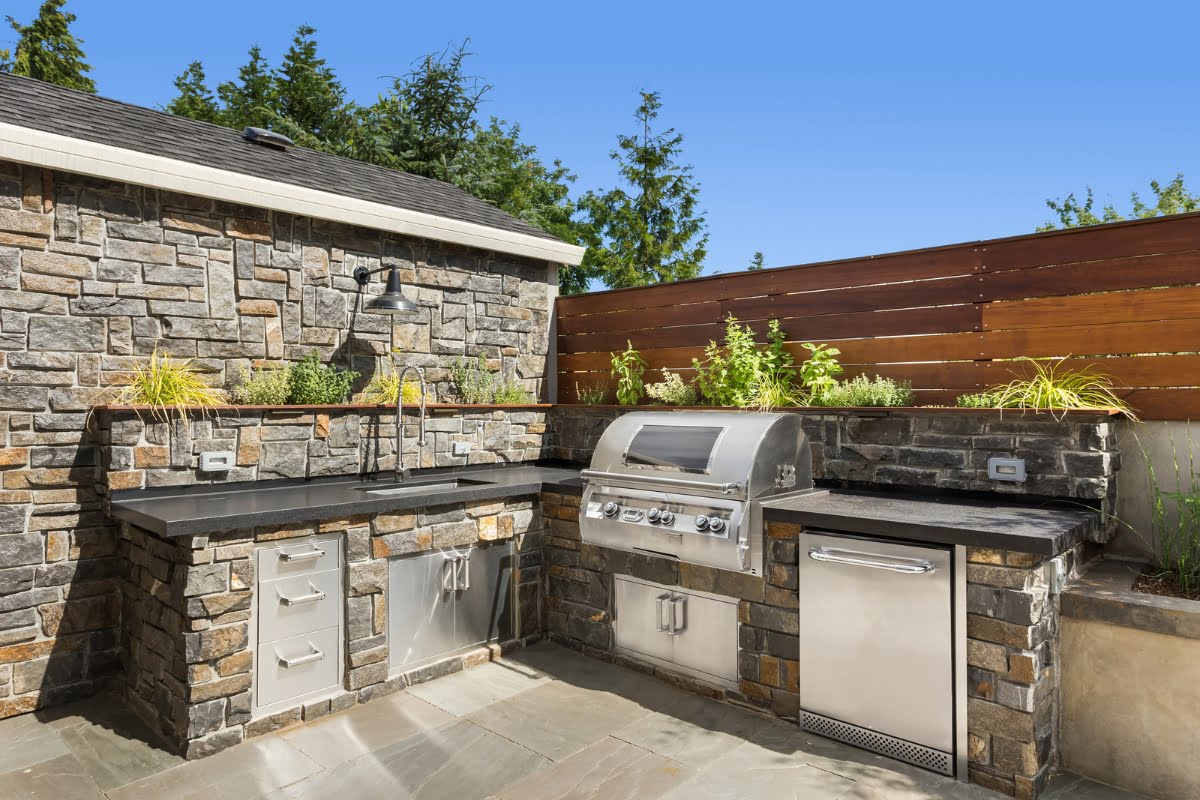 Outdoor kitchen designs come to life with stone countertops, a stainless steel barbecue grill, sink, and storage cabinets, all set against a stone wall and wooden fence.