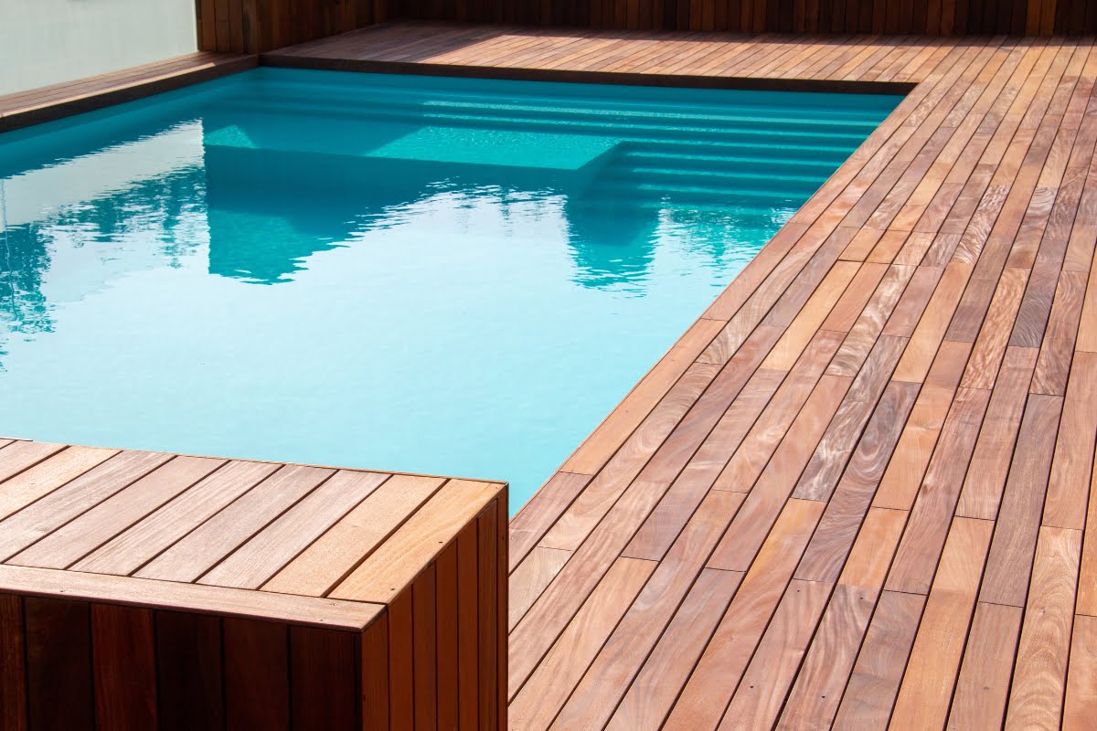 Rectangular swimming pool with clear blue water, surrounded by a wooden patio decking material. Steps lead into the pool at the far end.