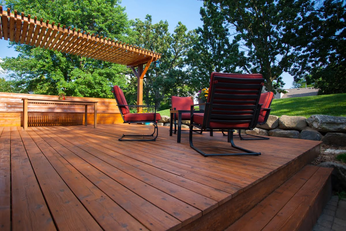 A wooden patio with a pergola, red cushioned chairs, a table, and surrounding trees on a sunny day, showcasing high-quality patio decking material.