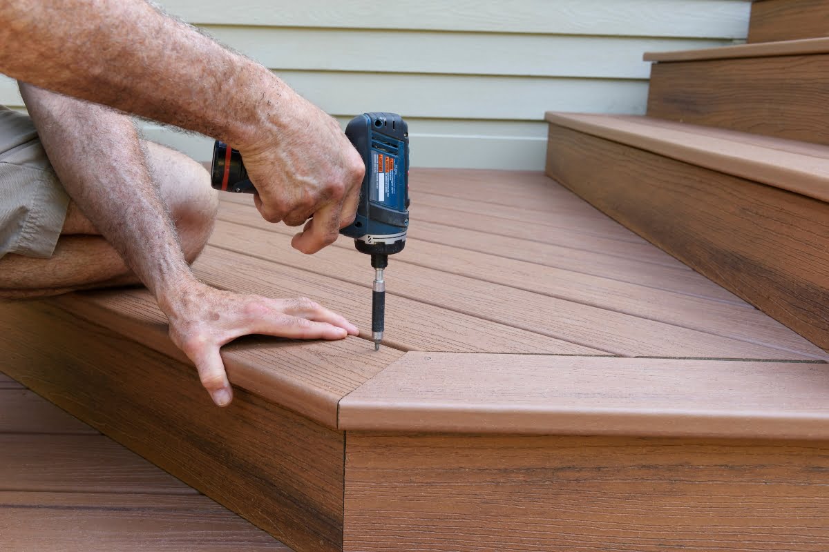 A person using a power drill to screw a brown wooden plank on an outdoor staircase made with patio decking material. Various wooden steps and a portion of a wall are visible.