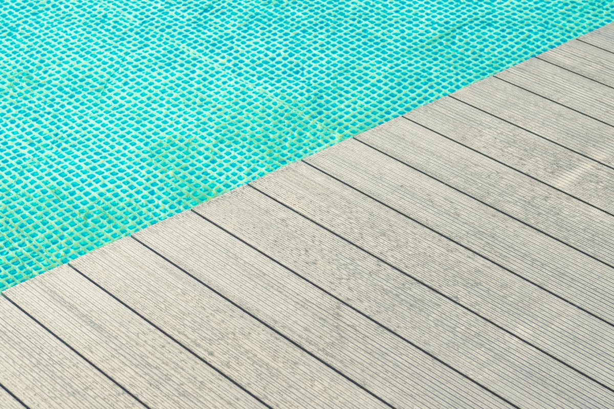 A close-up view of a swimming pool's edge with blue tiles beside a gray wooden deck, highlighting the sleek patio decking material.