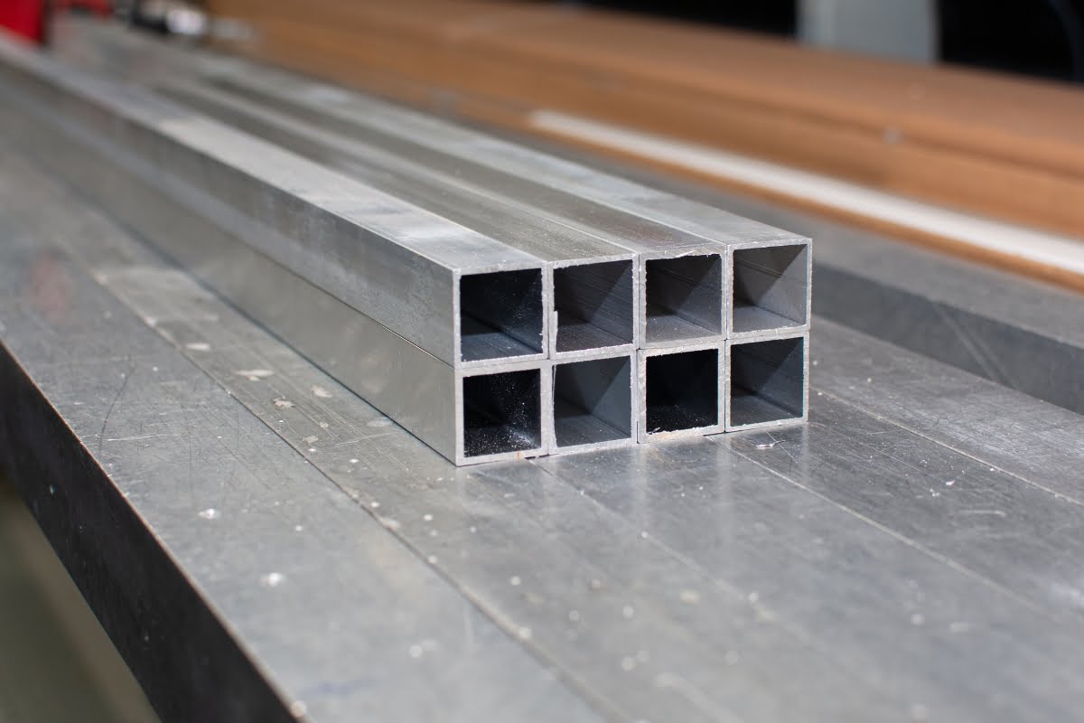 A stack of six hollow, square aluminum tubes, ideal for patio decking material, is placed on a metal surface.