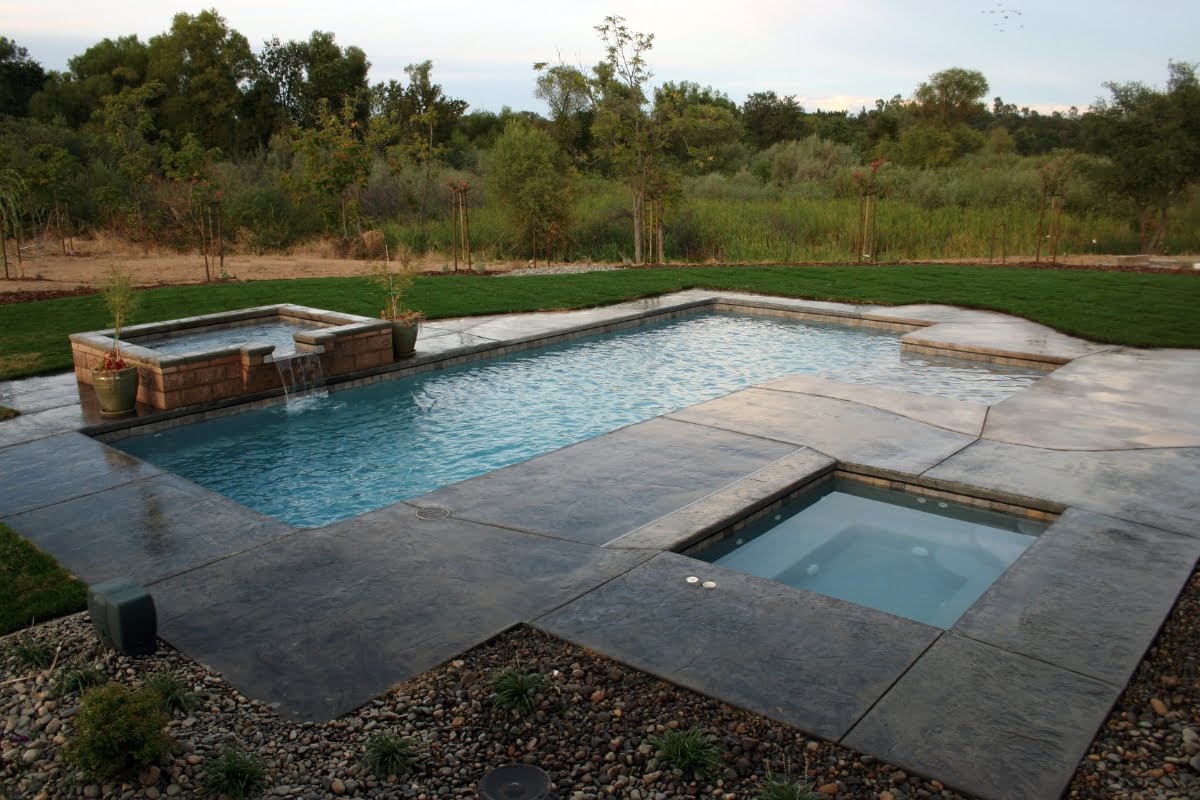 Outdoor pool area with a large rectangular pool, adjacent hot tub, and surrounding paved patio decking material. Green lawn and trees in the background.