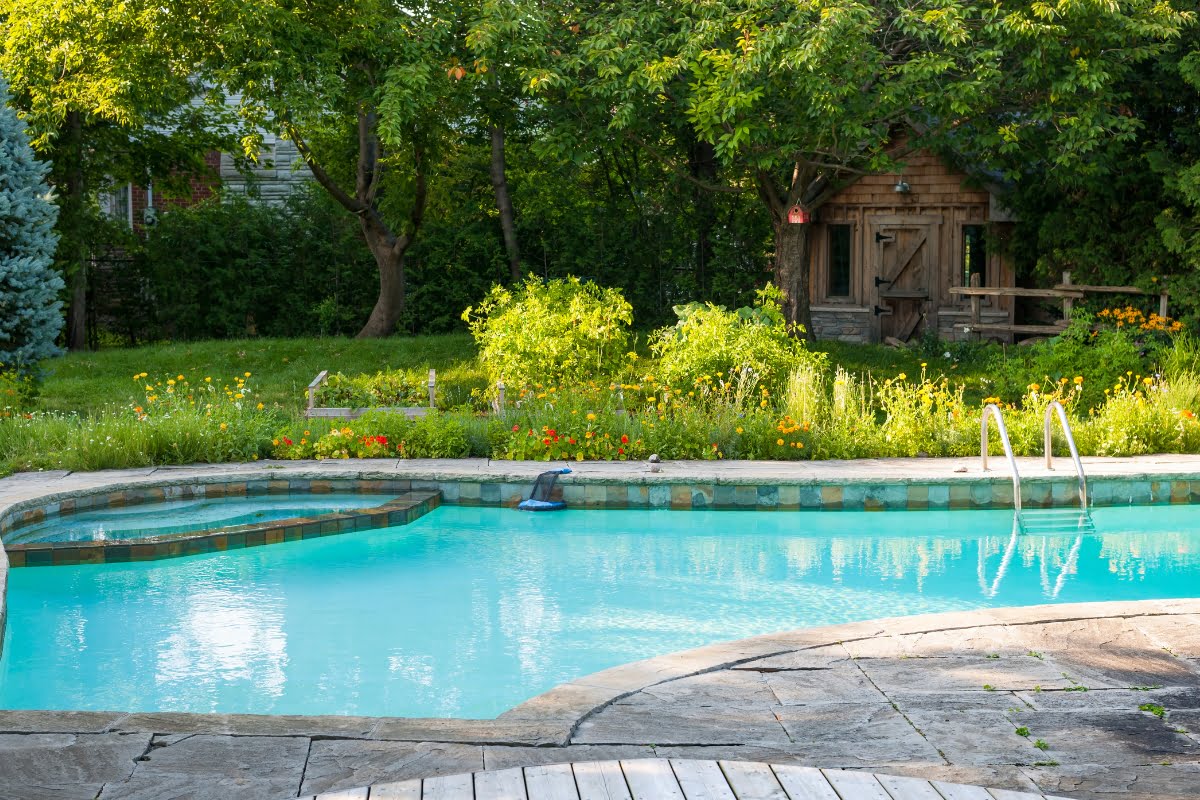 A clear swimming pool with steps and a ladder is surrounded by green trees and a garden, with patio decking material enhancing the area. In the background, there’s a small wooden structure adding charm to the serene setting.