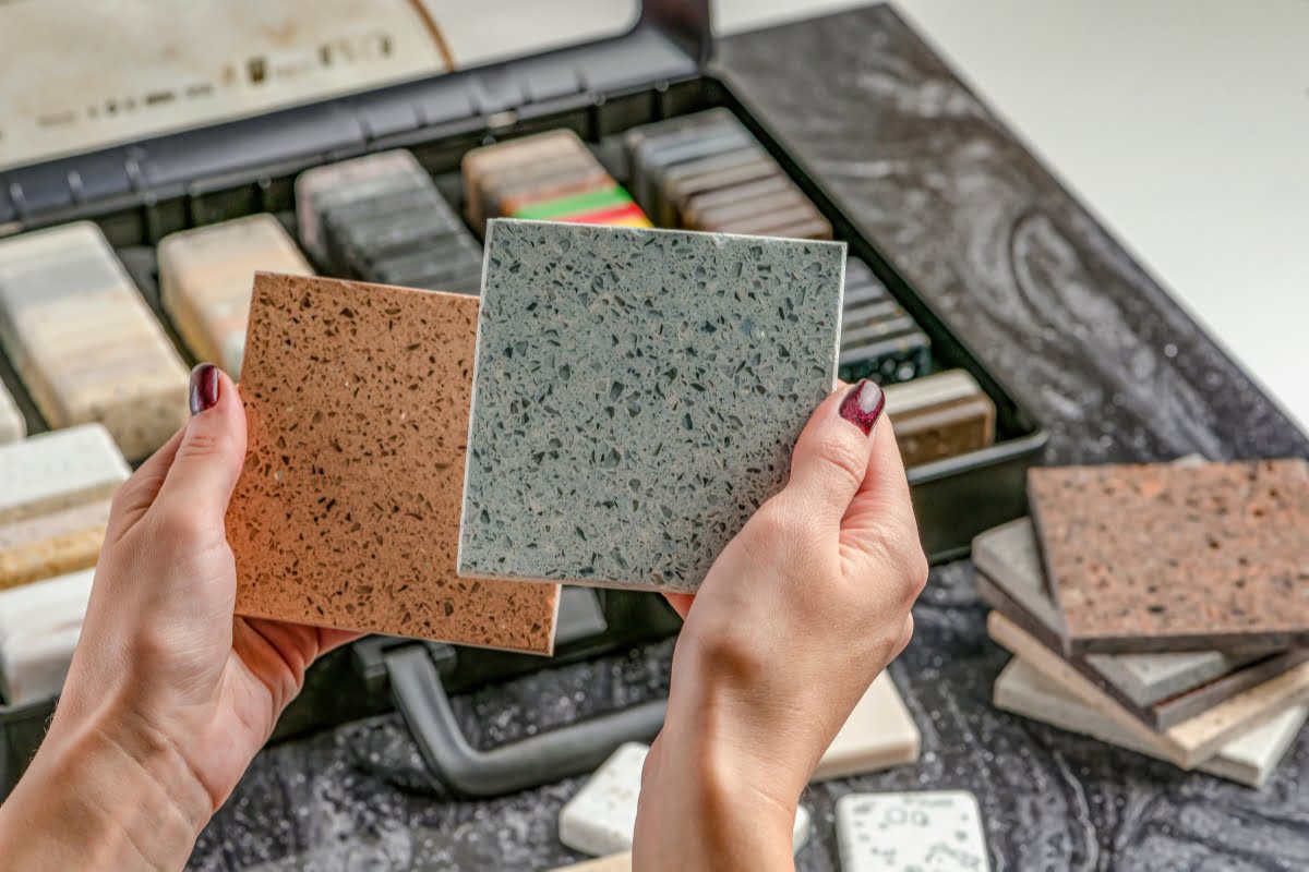 Hands holding two different stone countertop samples, one brown and the other grey, with a display box of various patio decking materials and samples in the background.