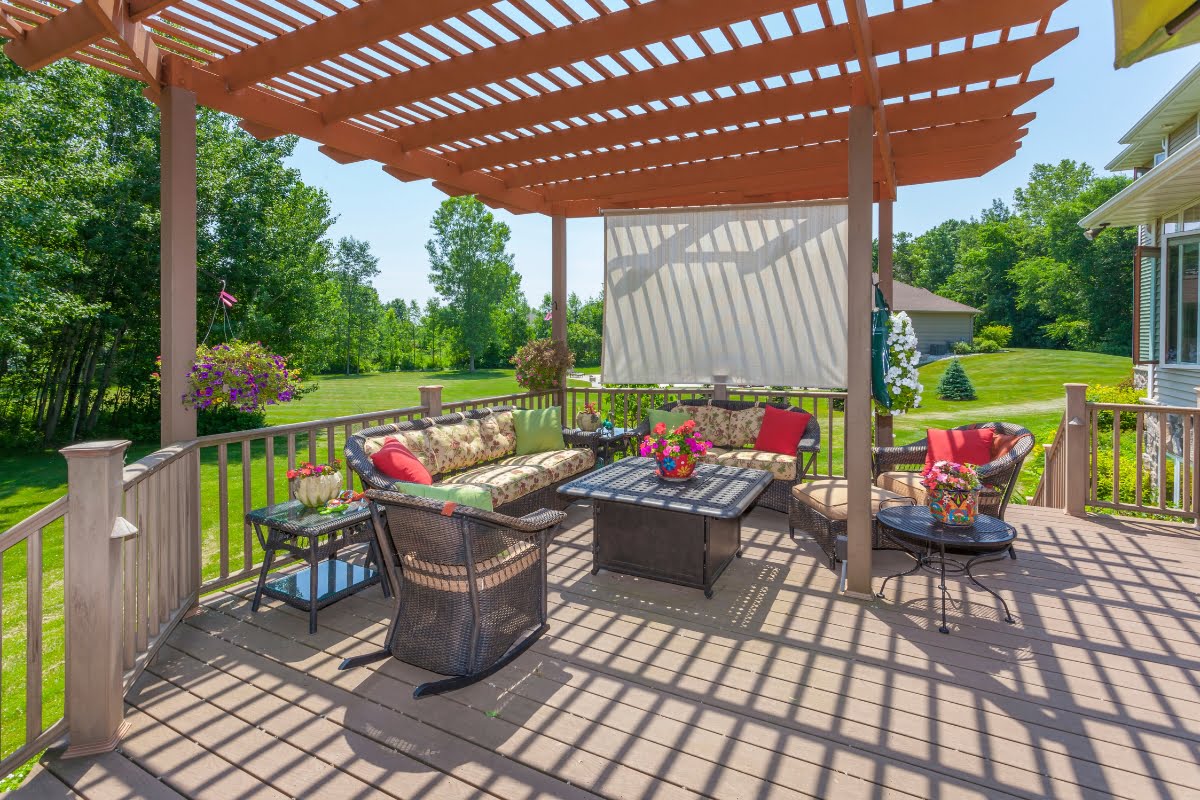 A backyard patio with a wooden pergola, comfortable seating, a patterned rug, and a table. The patio decking material complements the outdoor space beautifully, while flowers and greenery surround the area.