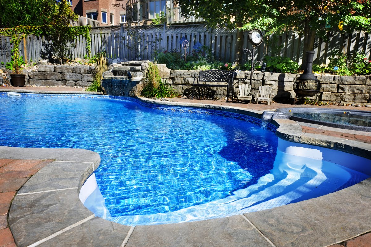 Backyard swimming pool with clear blue water, stone border, adjacent hot tub, landscaped garden, and seating area in the background—perfect inspiration for those seeking pool renovation tips.