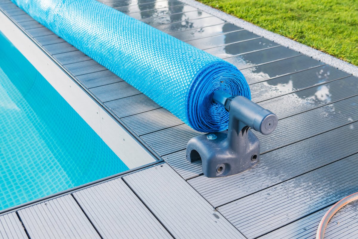 Close-up of a rolled-up blue pool cover on a reel system, positioned beside a clean, rectangular swimming pool with a tiled deck and adjacent green lawn—perfect inspiration for those seeking practical pool renovation tips.