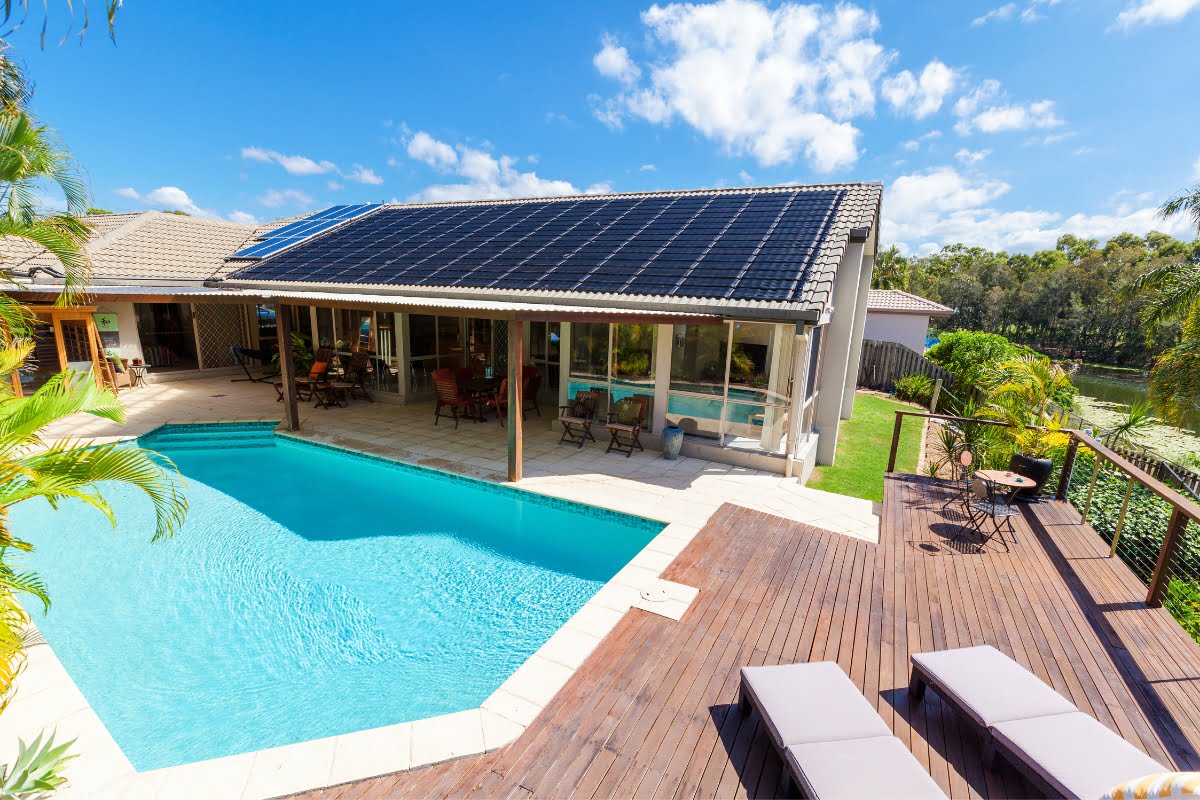 A modern house with solar panels on the roof, featuring a swimming pool that showcases recent pool renovation tips, a wooden deck with lounge chairs, and surrounding greenery under a clear blue sky.