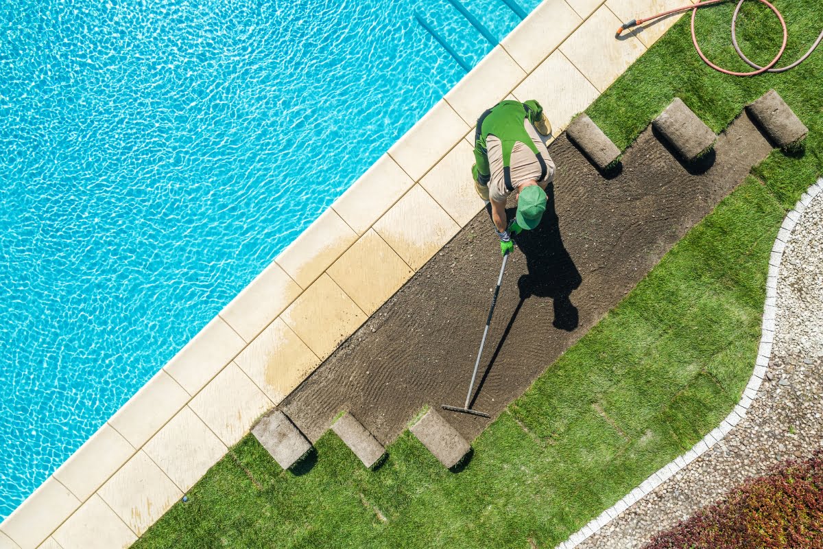 A person wearing green clothing is raking soil next to a swimming pool, preparing to lay down new sod pieces. The pool, surrounded by paving stones and a grassy area, seems poised for an upgrade—an ideal scenario to explore some valuable pool renovation tips.