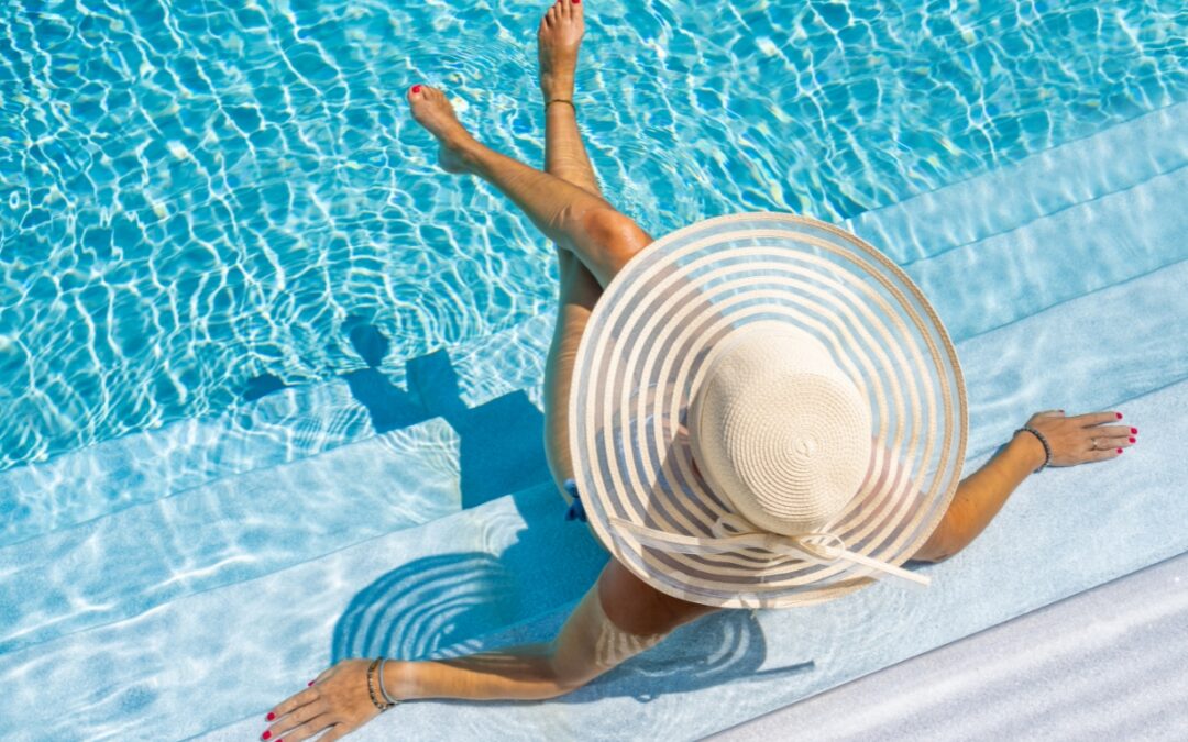 A person with painted nails sits on the edge of a pool spa combo, wearing a large, wide-brimmed hat that creates a striped shadow pattern on the water.