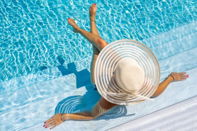 Person wearing a wide-brimmed sun hat sits on pool spa combo steps with legs crossed, partially in the water, under bright sunlight.