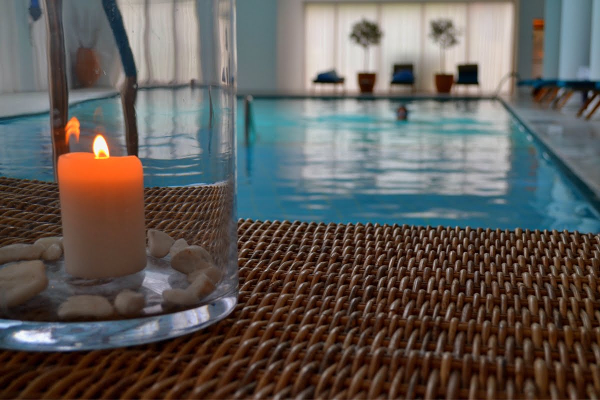 A lit candle in a glass holder sits on a woven surface by an indoor pool spa combo, with a swimmer gracefully gliding in the background.