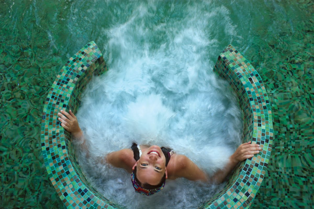 A person smiles while relaxing in a tiled, circular pool spa combo with jets creating bubbles around them. The surrounding water is clear and green.