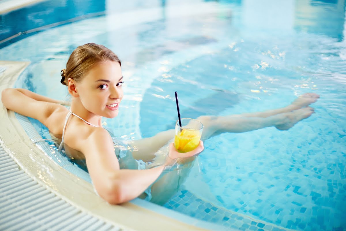 A woman indulges in a pool spa combo, relaxing as she holds a drink with a straw and an orange slice.