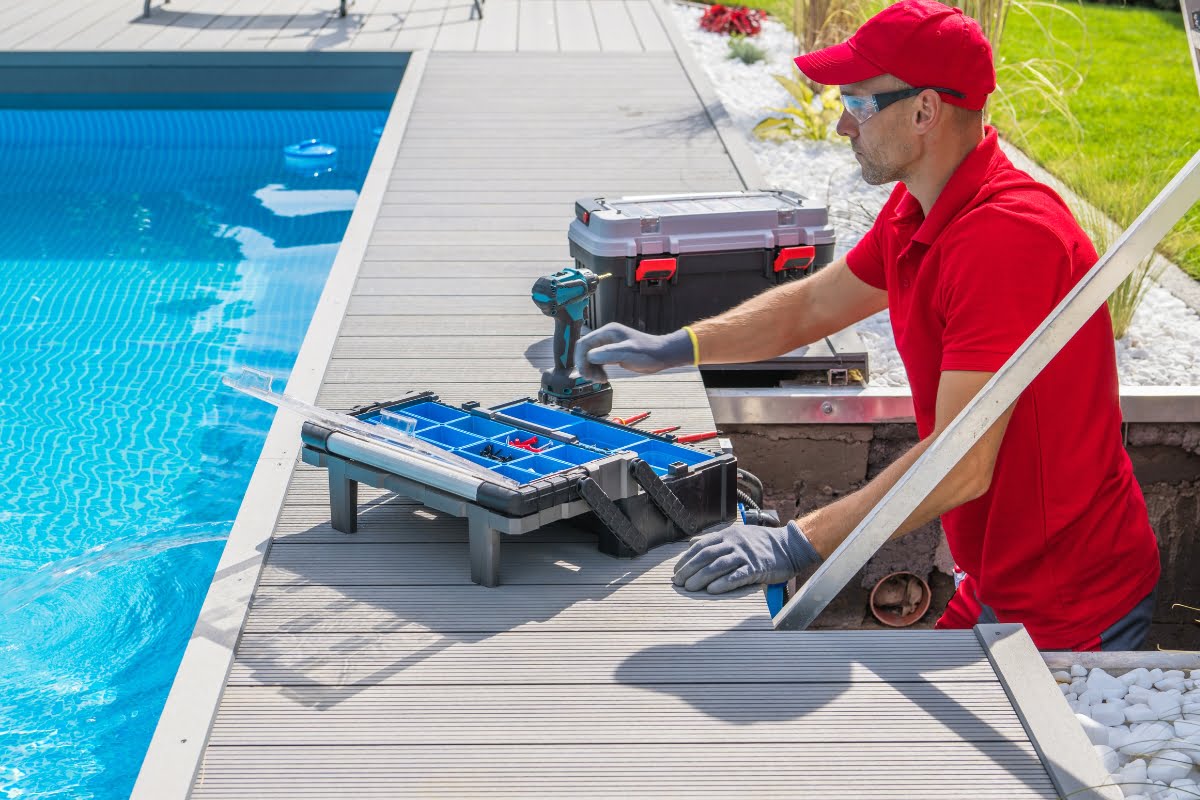 A man in a red shirt and cap works on pool maintenance for a pool spa combo. He uses tools from an open toolbox next to him while kneeling by the poolside. A drill and a closed toolbox are also on the deck.