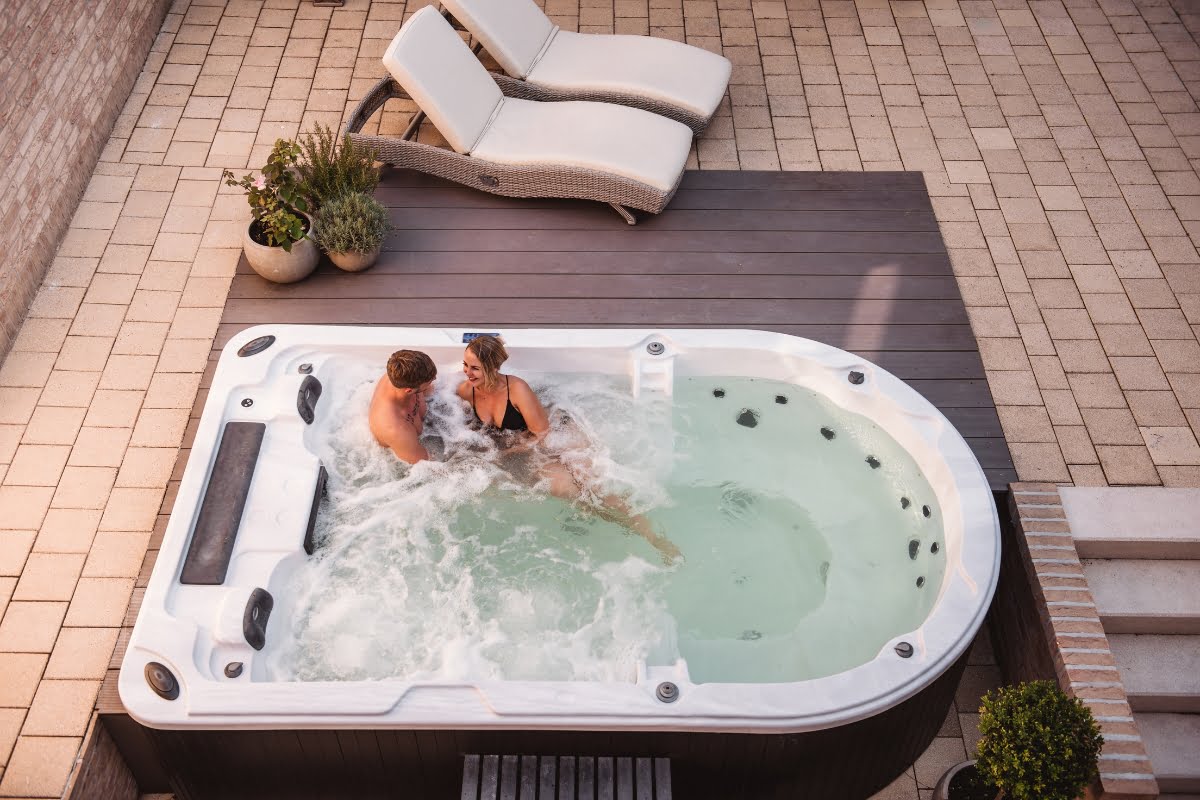 A couple relaxes in an outdoor pool spa combo with water jets on, surrounded by a paved patio. Two lounge chairs and potted plants are nearby, enhancing the serene atmosphere.