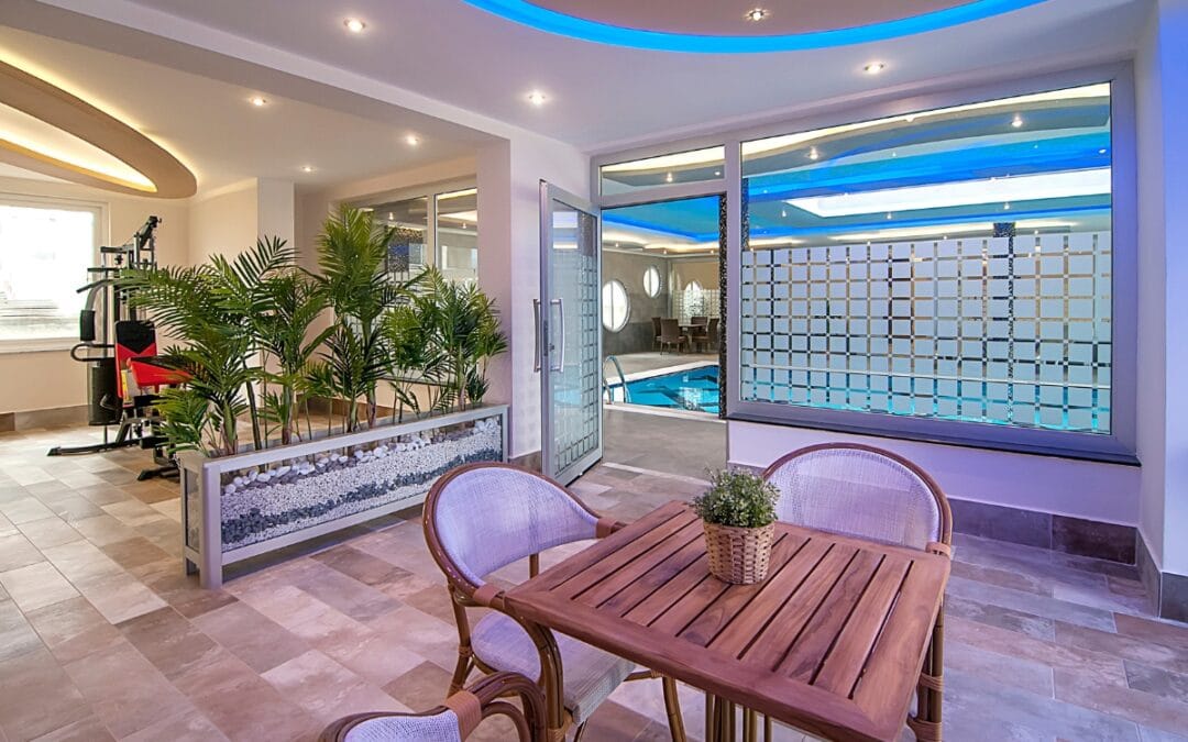 The indoor gym with pool features a seating area with a wooden table and chairs, situated adjacent to the water behind glass doors. The space is adorned with potted plants and enhanced by blue accent lighting on the ceiling.