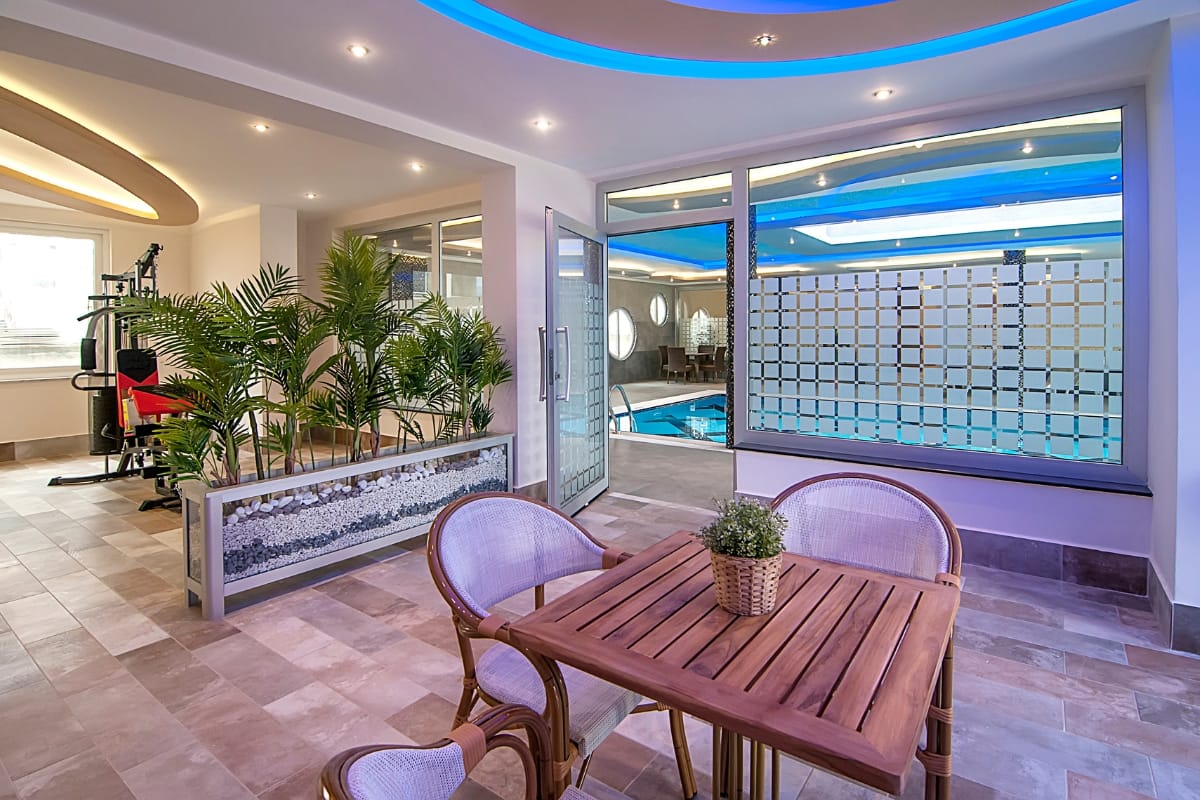 Modern indoor leisure area featuring a wooden table and chairs, potted plants, and large windows overlooking a gym with pool and fitness equipment.