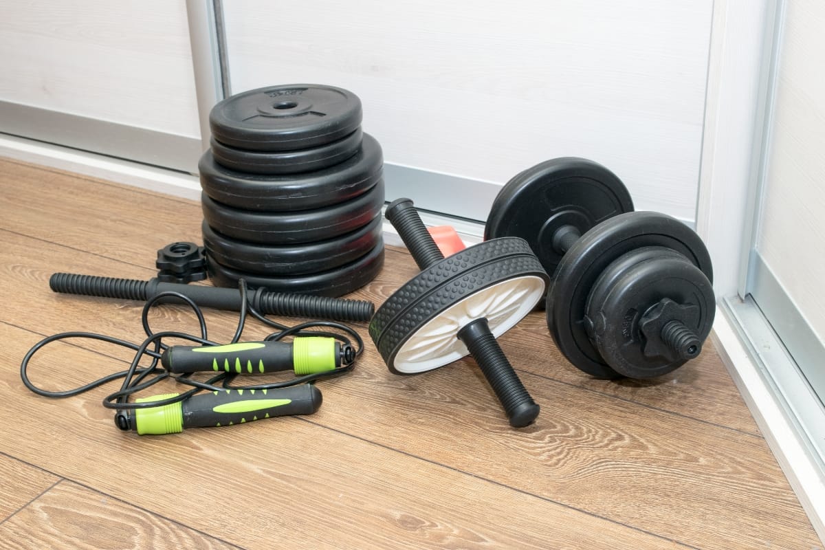 A set of adjustable dumbbells, weight plates, a jump rope, and an ab wheel are neatly arranged on the wooden floor, ready for a workout. Imagine this setup in a gym with a pool to unwind after your session.