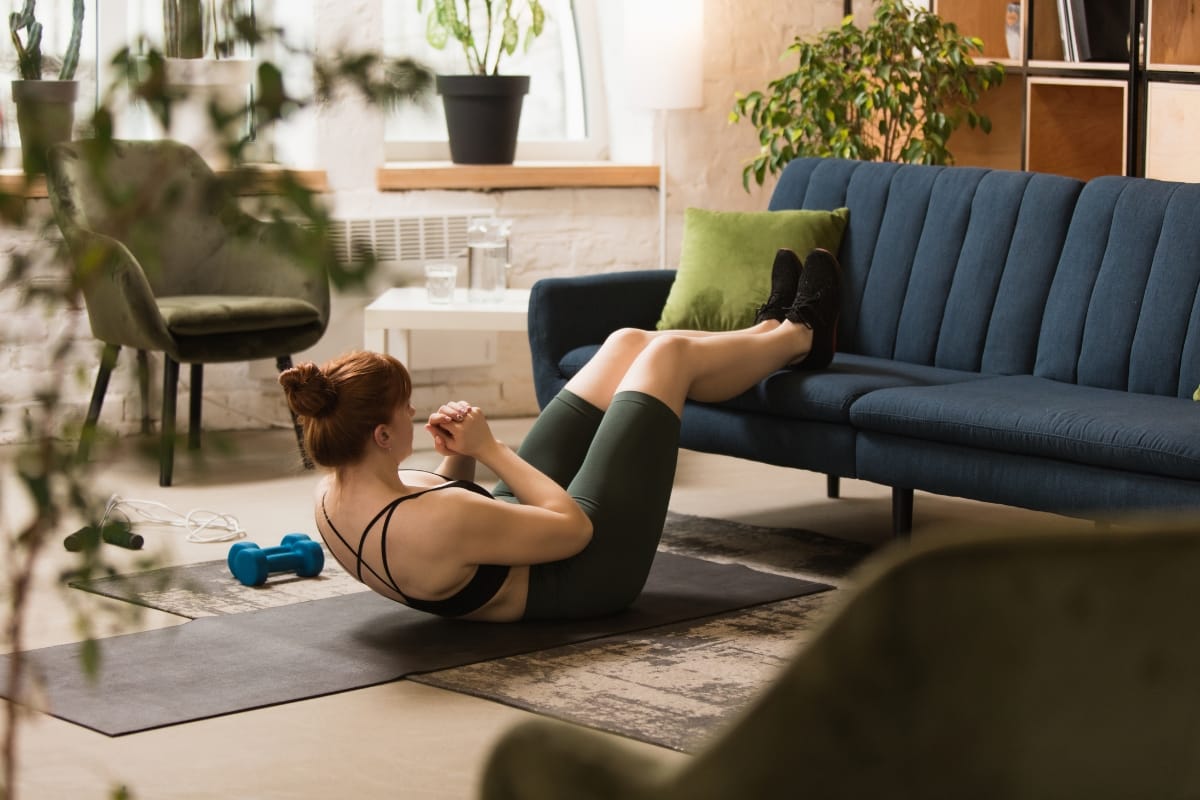 A person performs a crunch exercise on a mat in a cozy living room adorned with plants and complemented by a blue sofa, dreaming of their next session at the gym with pool facilities.