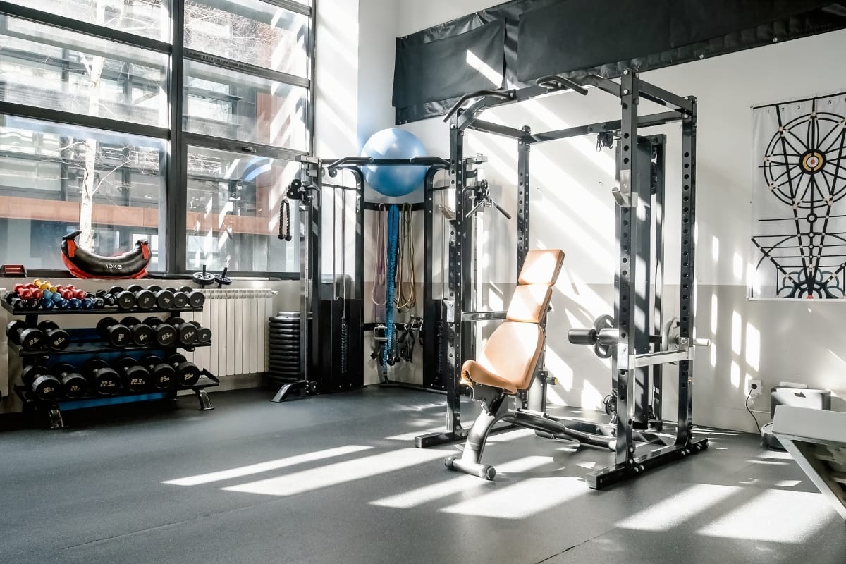 A sunlit gym featuring a weight bench, dumbbell rack, resistance bands, and exercise ball, with the added luxury of a refreshing pool.