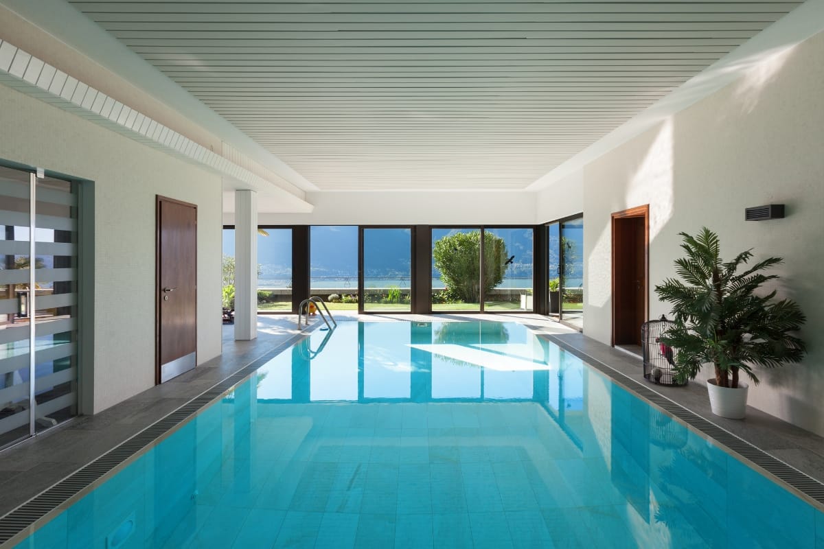 Indoor swimming pool with clear water, glass doors, and a view of mountains and greenery. This gym with a pool features a ceiling with parallel lines, and a potted plant graces the right side.