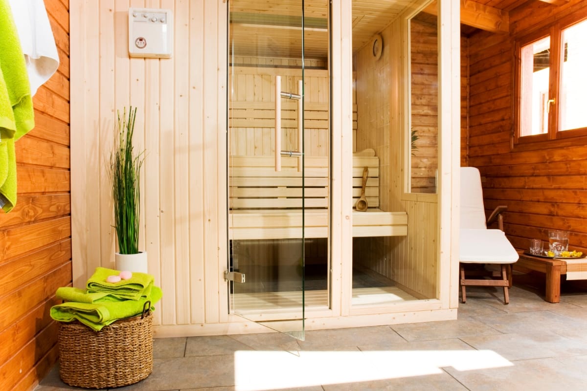 A wooden sauna room with a glass door overlooks the gym with pool. Inside, a bench and a rolled towel are visible. Outside, there's a wicker basket with towels and a lounge chair beside a small table, inviting relaxation after your swim.