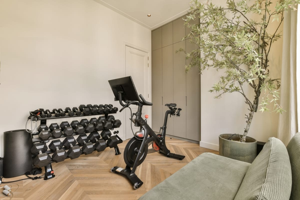 A home gym with a stationary bike, a rack of dumbbells, and a large plant seamlessly complements the room's wood flooring and neutral-colored walls, offering a tranquil workout space that feels as refreshing as a gym with pool amenities.