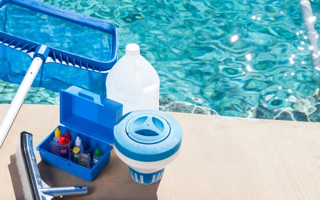 Pool cleaning essentials for winterizing your pool, including a net, water testing kit, liquid container, and floating chlorine dispenser, are placed on the edge of a swimming pool.