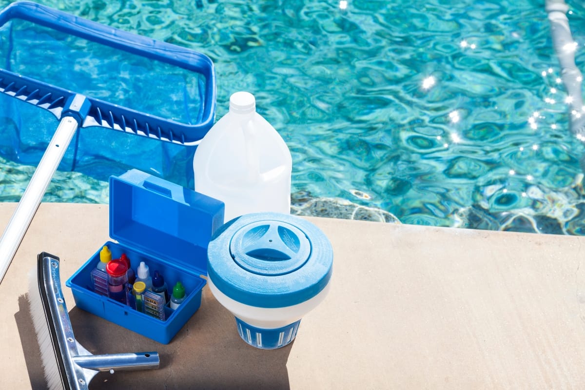 Pool cleaning supplies, perfect for winterizing your pool, including a net, water testing kit, gallon container, and floating dispenser, are arranged on the poolside next to clear blue water.