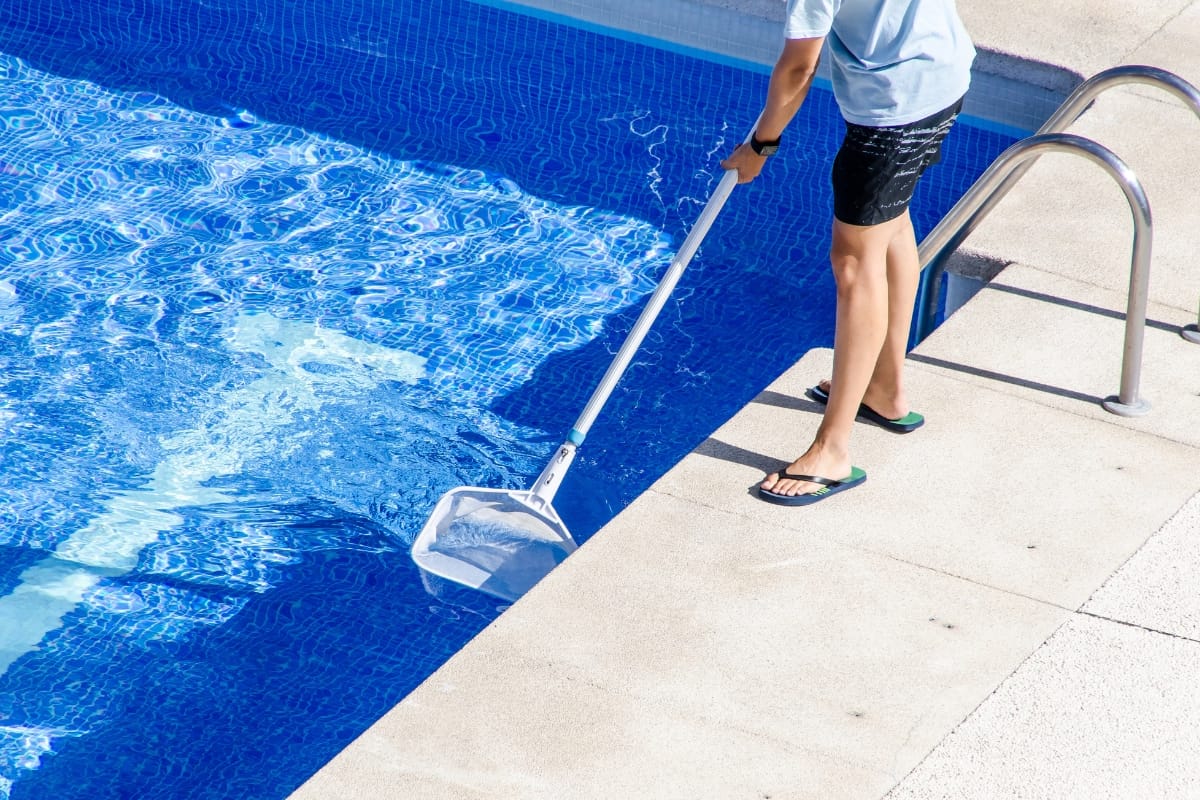 Under the sun's warm embrace, a person diligently cleans the swimming pool with a skimming net, all in preparation for winterizing your pool.