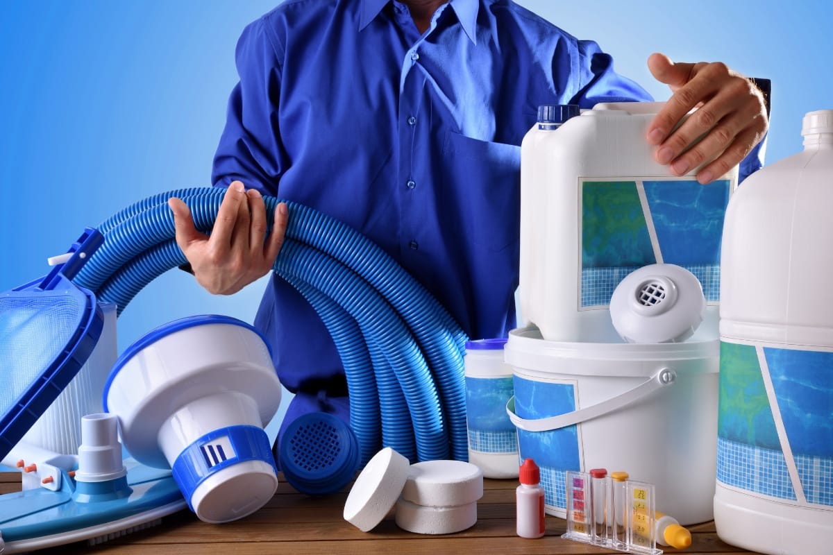 Person in blue shirt holding pool cleaning equipment and chemicals, including hoses, containers, and test kits for winterizing your pool, arranged on a wooden surface with a blue background.