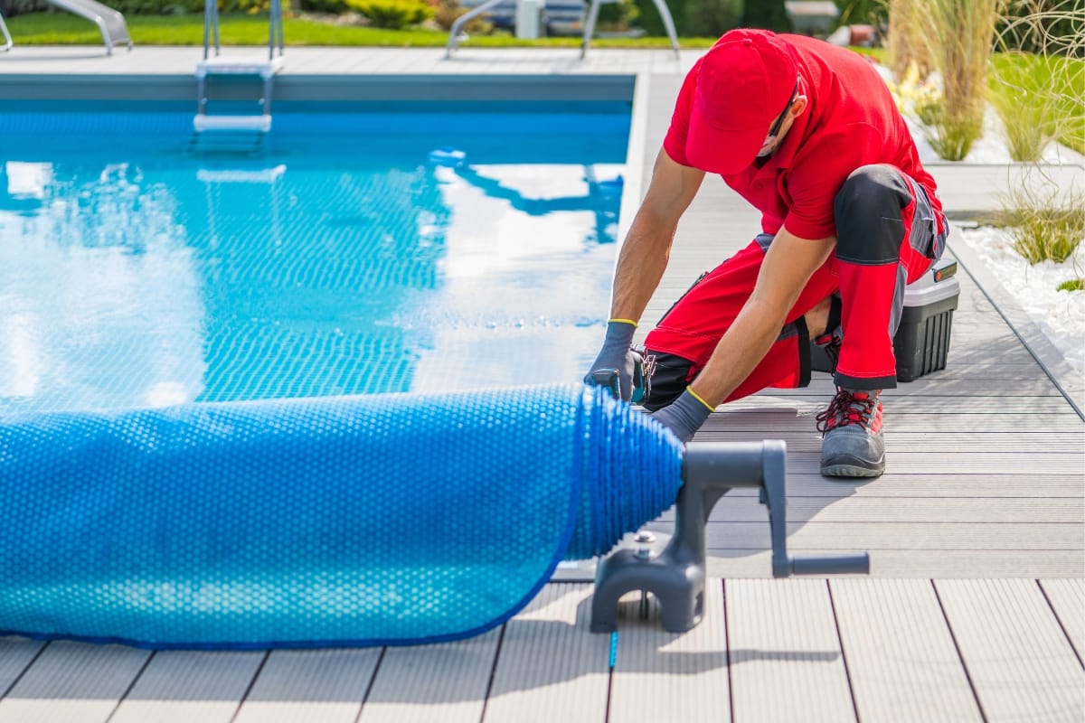 A worker in red uniform is kneeling and rolling up a blue pool cover on a bright, sunny day, marking the first step in winterizing your pool.