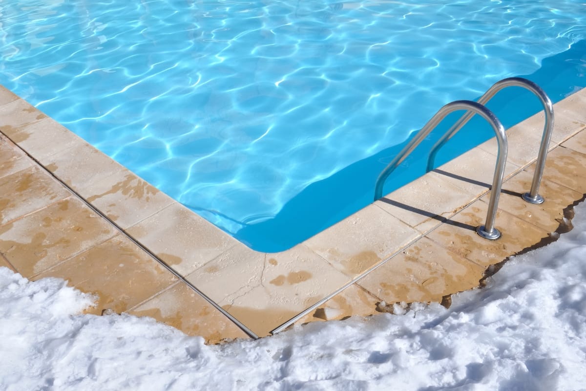 A swimming pool with clear blue water is partially surrounded by snow, a serene winter scene. As the season changes, consider winterizing your pool to maintain its pristine condition. A metal pool ladder is visible on the right side.