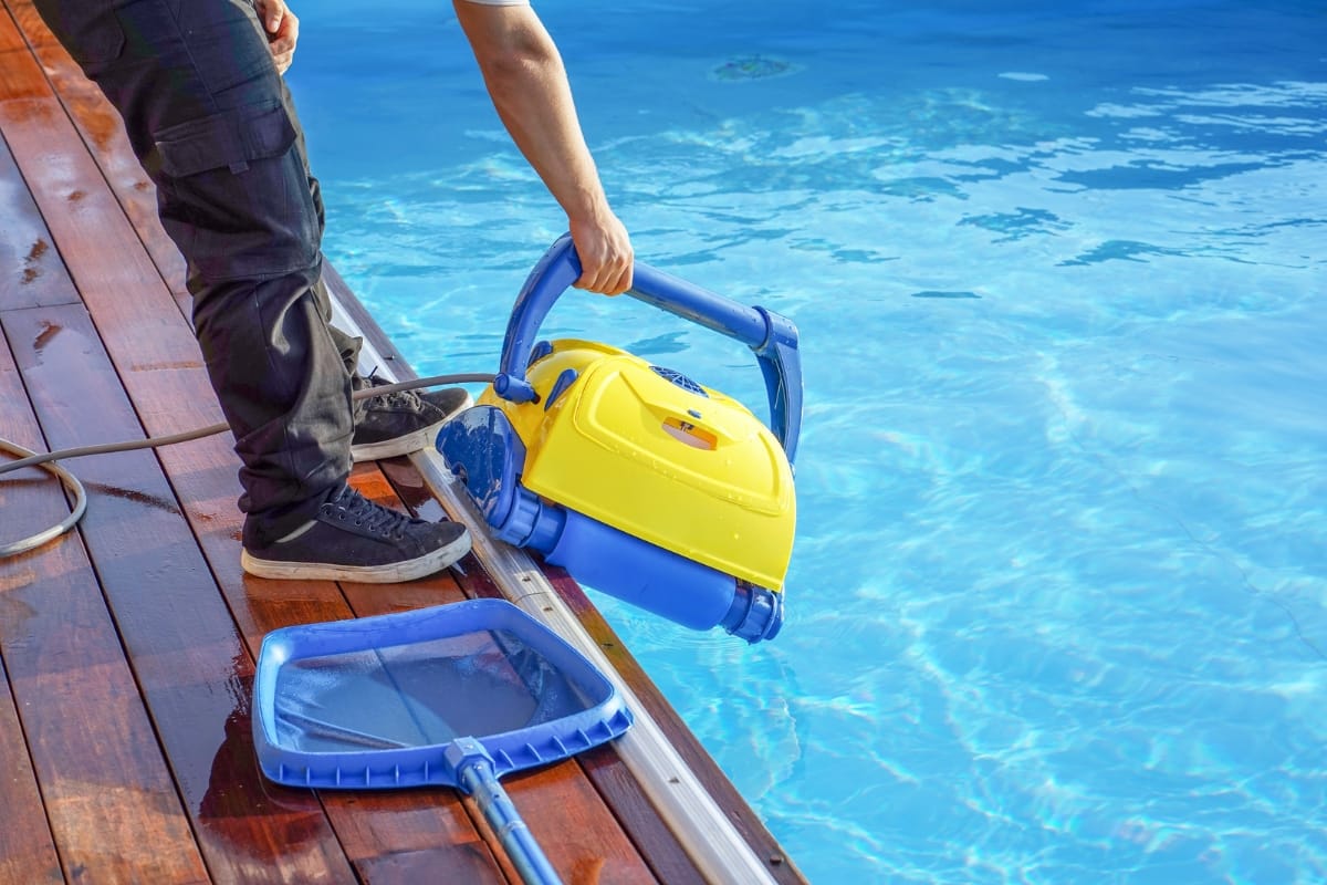 A person places a yellow and blue robotic pool cleaner in the swimming pool, preparing for winterizing your pool, with a net and hose nearby on the wooden deck.