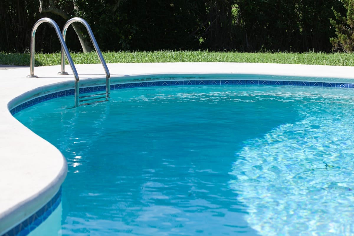 A clear, blue swimming pool with metal ladder steps and a curved white concrete border sits next to a grass lawn, ready for winterizing your pool when the season changes.